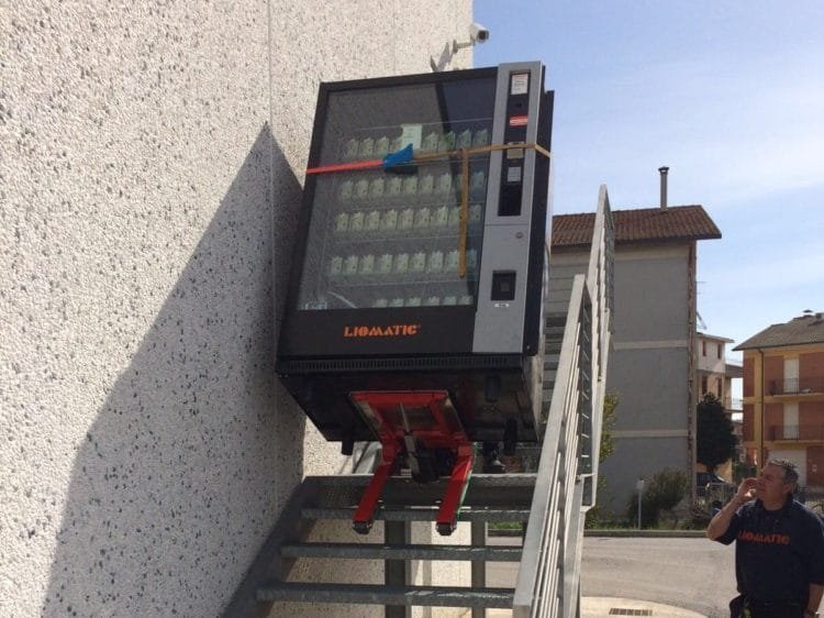 Un chariot monte-escalier électrique permet de transporter un distributeur de boissons lourd sur un escalier métallique externe.